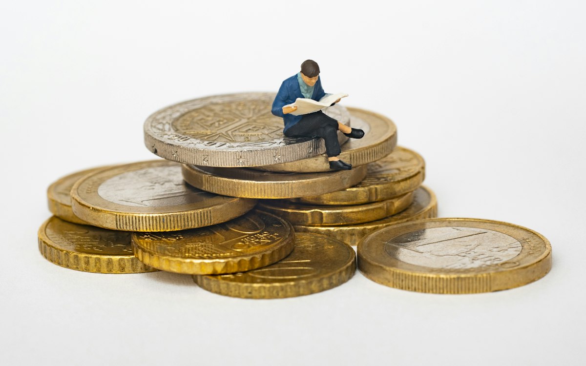Miniaturfigur eines lesenden Geschäftsmanns auf einem Stapel Euromünzen – Liquidität durch vorausschauendes Forderungsmanagement sichern